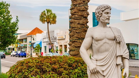 An image of a St. Armands Circle Statue of a bearded man wearing a toga next to the base of a palm tree and a bush.